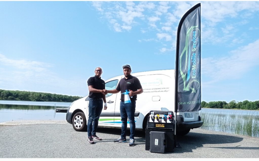 Deux membres de l'équipe EcoMobile serrant la main devant leur véhicule de service près d'un lac, avec du matériel de nettoyage écologique exposé et une bannière publicitaire